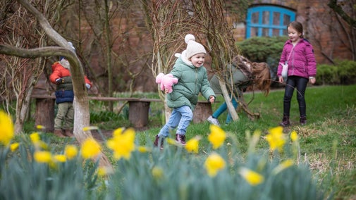 Child running, holding a teddy bear, with a background of trees and daffodils in the front.